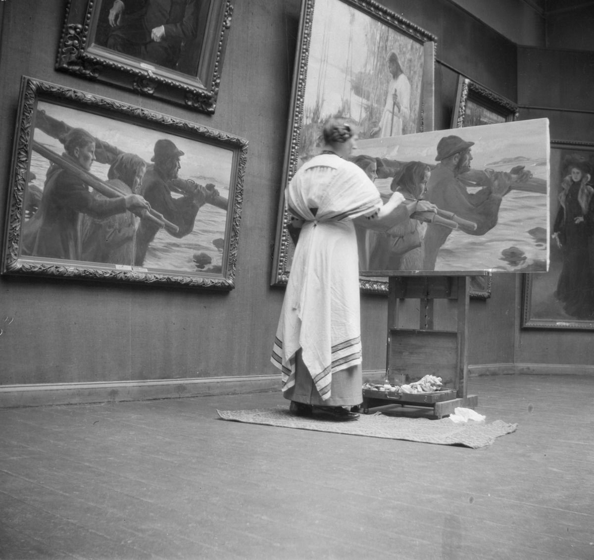 a black and white photograph of an exhibition hall in a museum. Multiple large paintings hang on a wall. A woman in a long dress stands in front of a canvas on an easel, she's painting a copy of one of the paintings that hangs on the wall next to her.