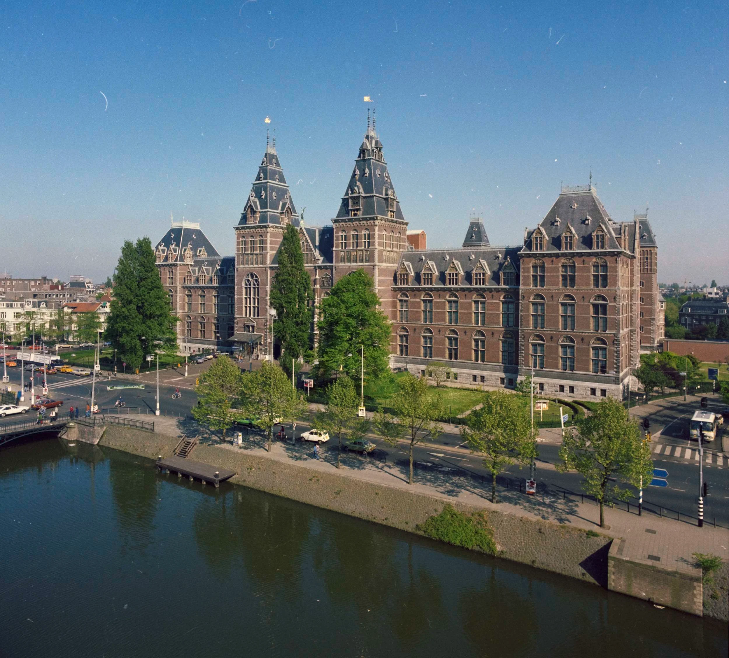 Luchtfoto van het Rijksmuseum met een gracht op de voorgrond, tegen een strakblauwe lucht.
