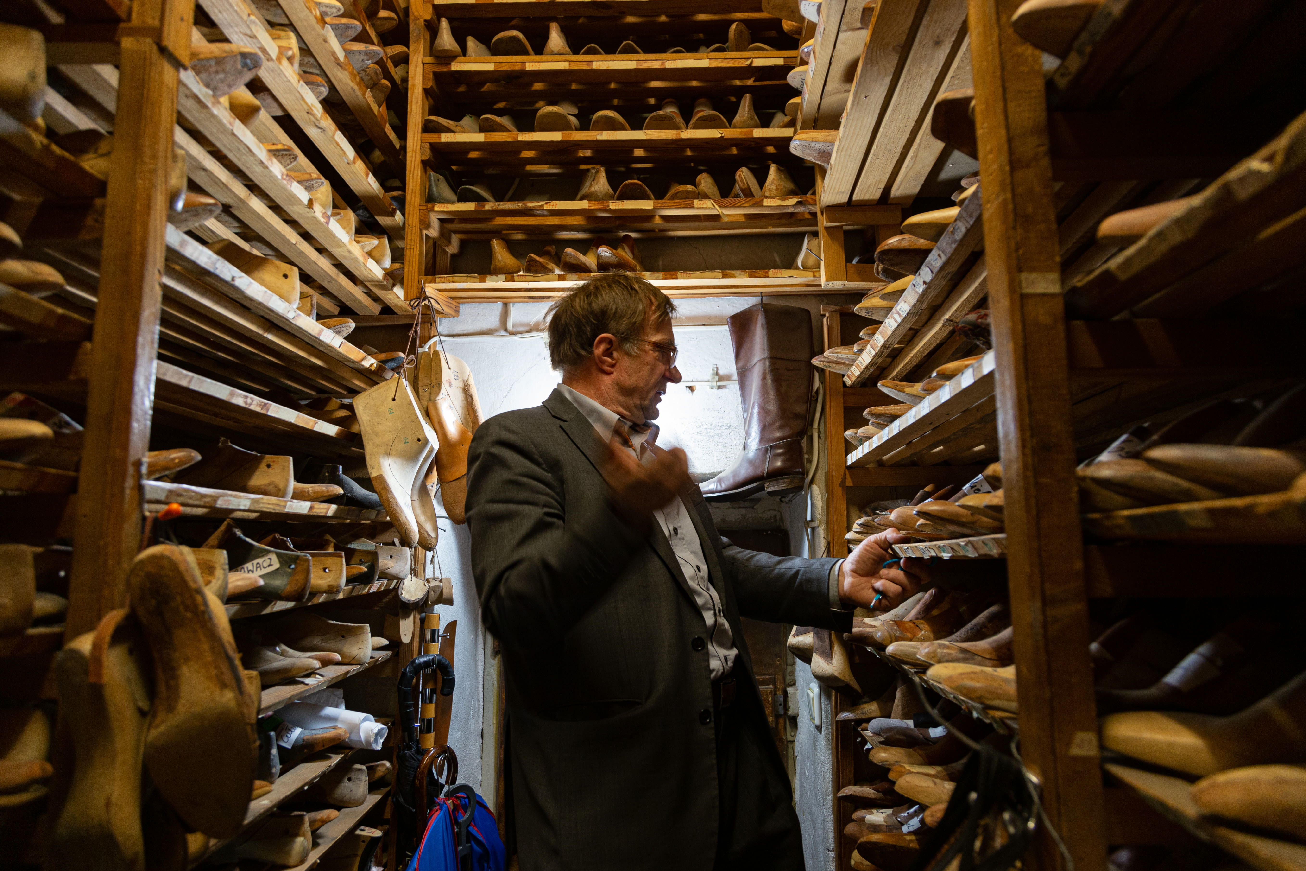 Jackek Kaminski, a Polish present-day shoe artisan, stand in a grey suit among shelves on either side and above him, all filled with brown wooden shoe molds.