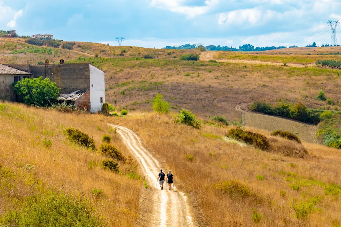 Two figures walking along a dust track that passes through dry fields. On the left hand side are square buildings, with electricity pylons visible in the distance. ©oltrelautostrada / Shutterstock.