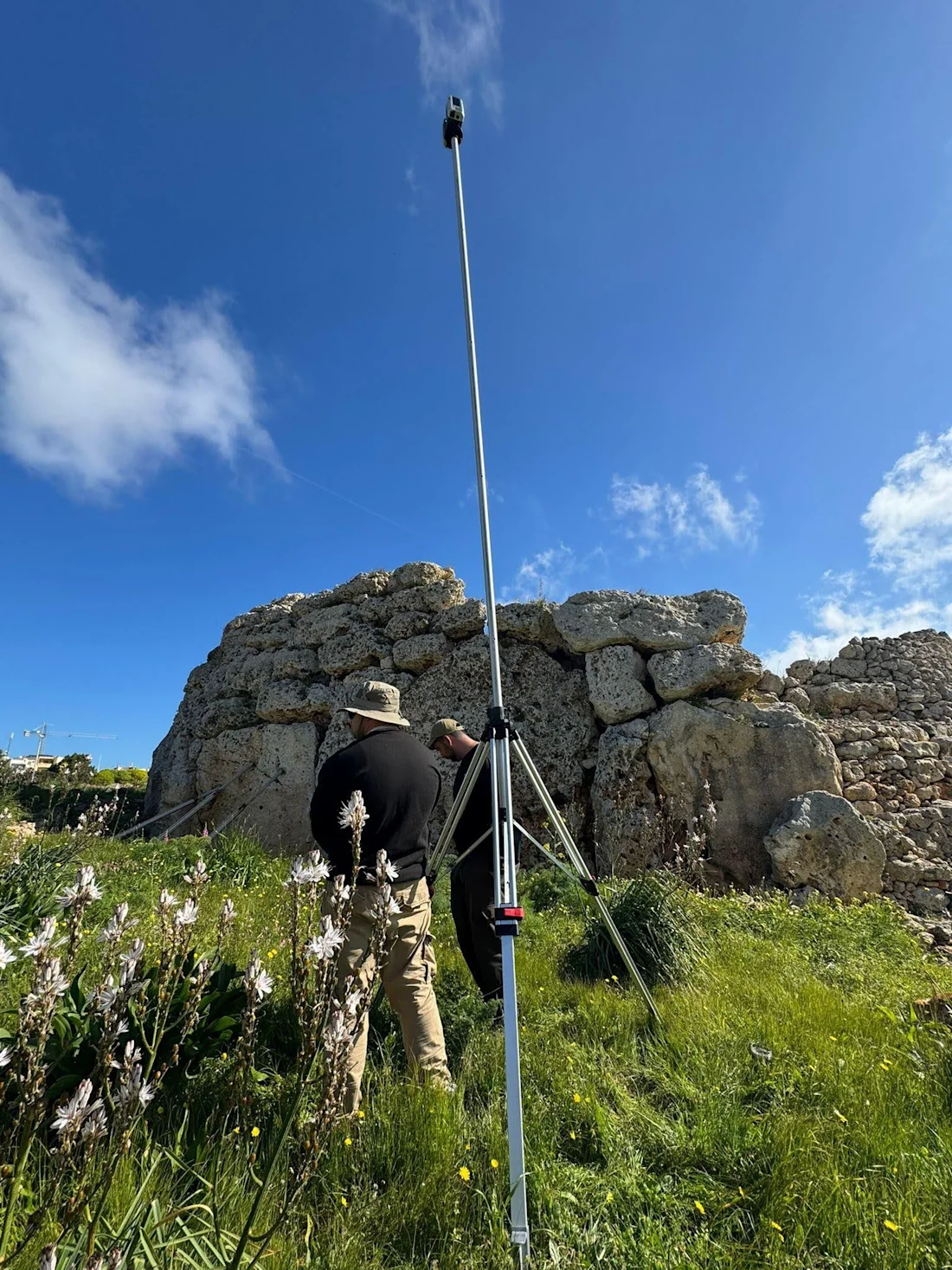 Person in a hat using surveying equipment near ancient stone ruins under a bright blue sky.