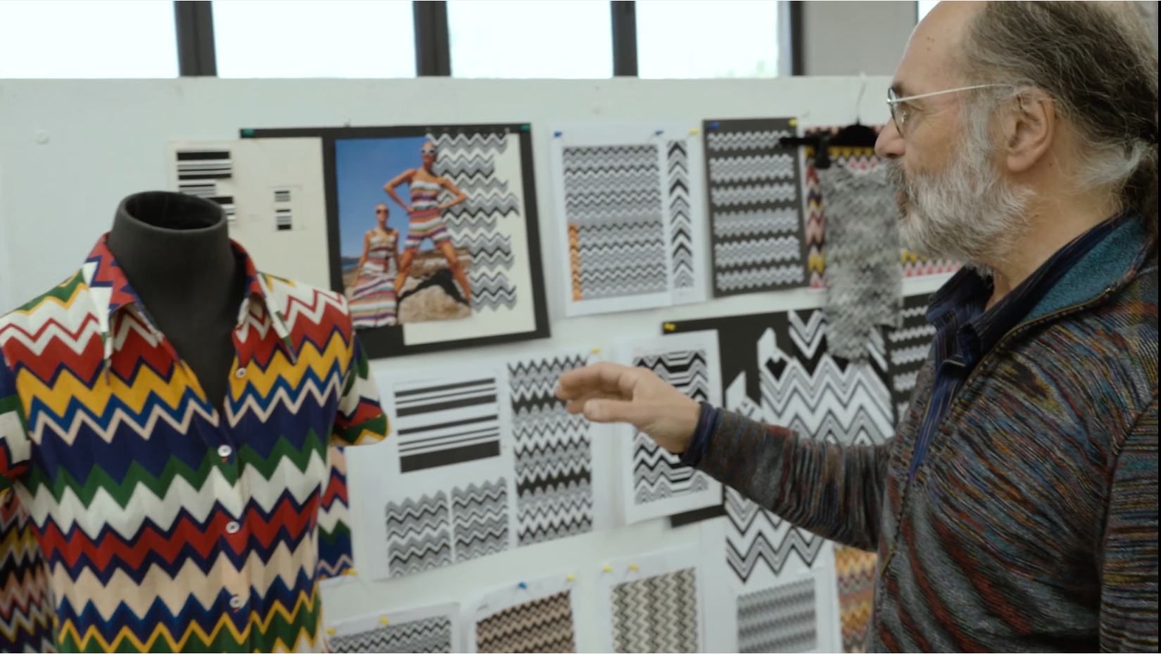 Luca Missoni stands in front of a design board with different patterns. On the left stands a mannequin dressed in a colourful patterned shirt from the Missoni line