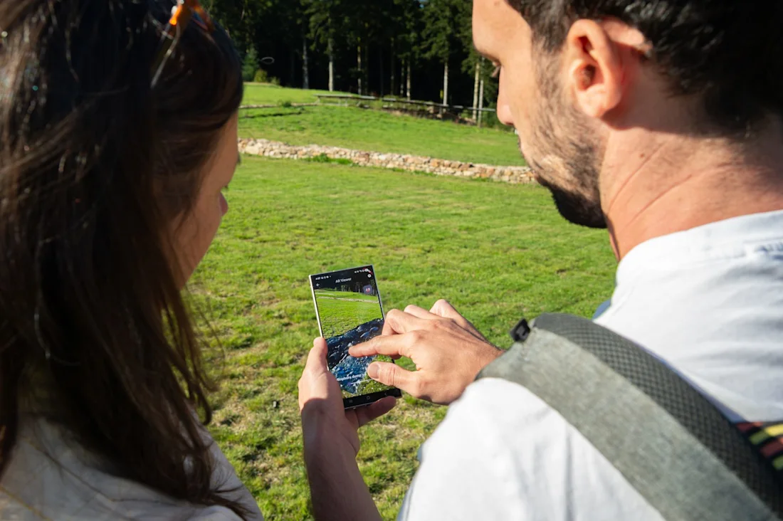 Two people outdoors look at a smartphone screen together in a grassy area with trees in the background.