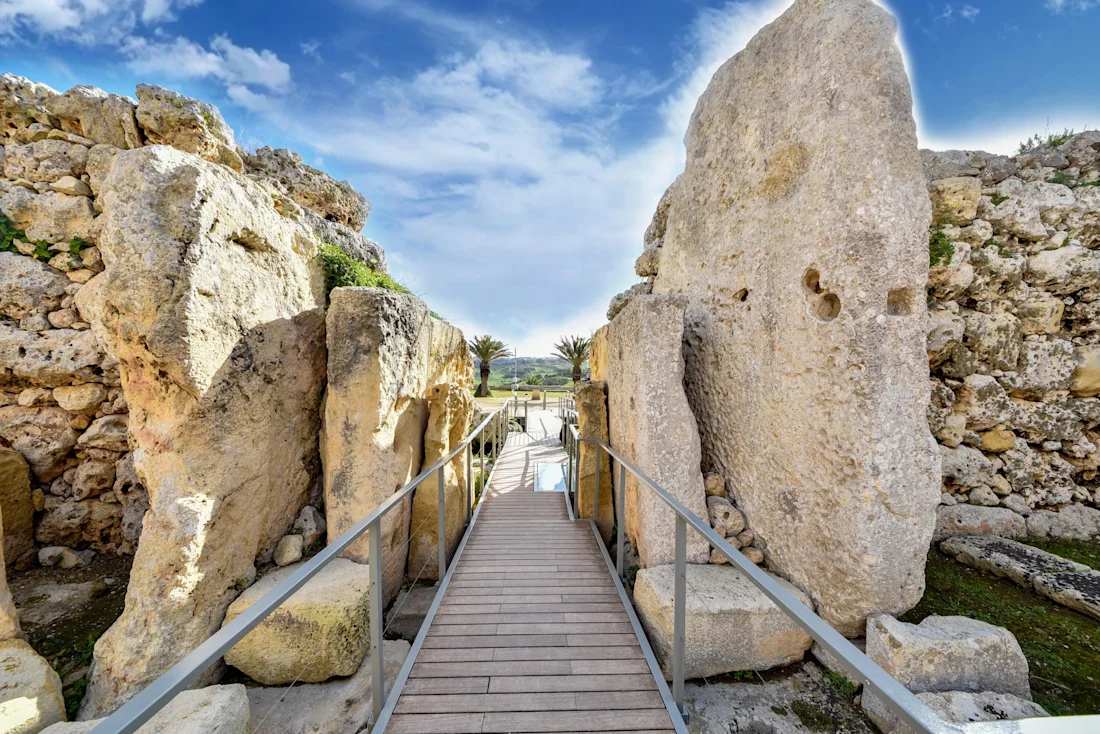 Stone pathway with railings leads through ancient large standing rocks under a partly cloudy blue sky.