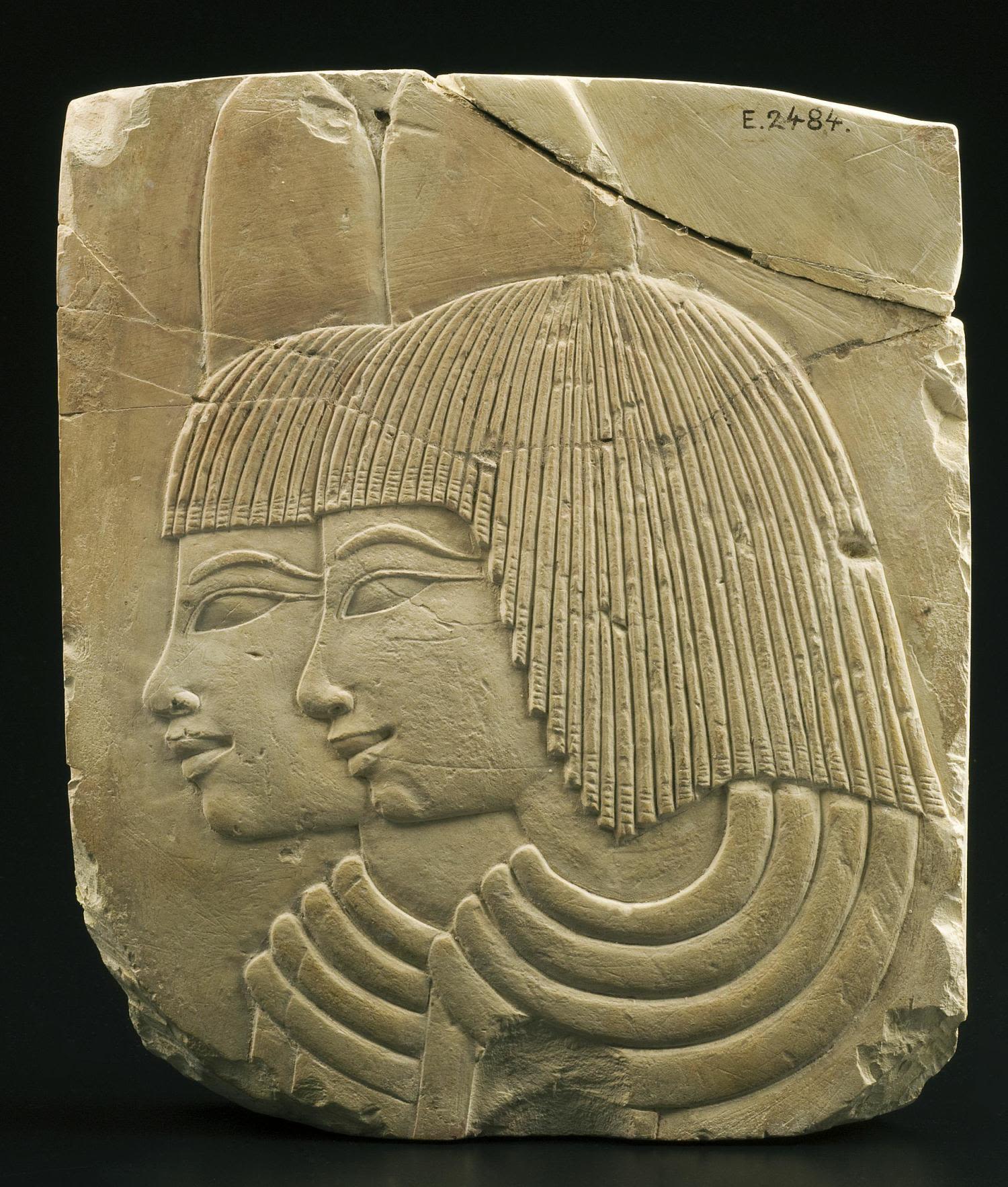 a gray stone slab on a black background with a relief sculpture of two female faces and necks pointing looking to the left, in Ancient Egyptian style