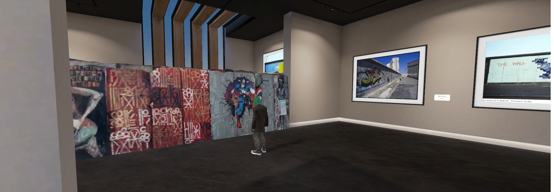 A figure stands in a museum, in front of a section of the Berlin Wall. Two photographs of the Wall are also exhibited.