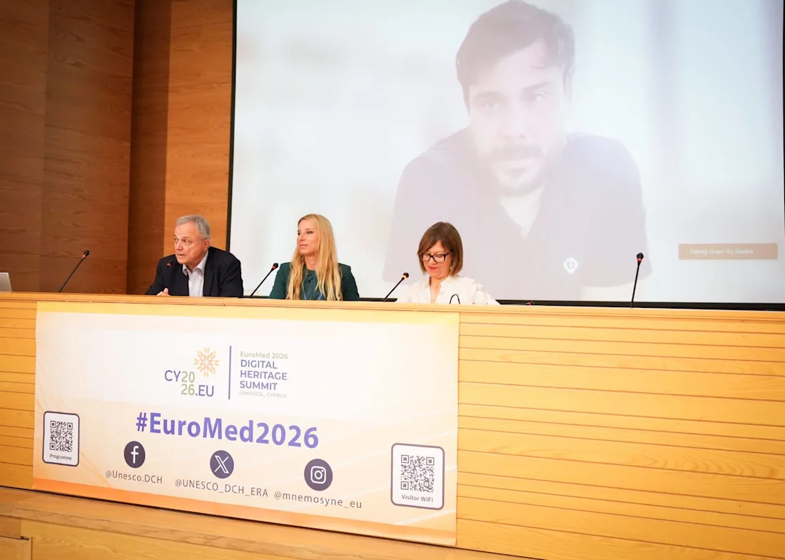Three people sit at a panel table with a large screen behind them showing a man’s face, at #EuroMed2026 event.
