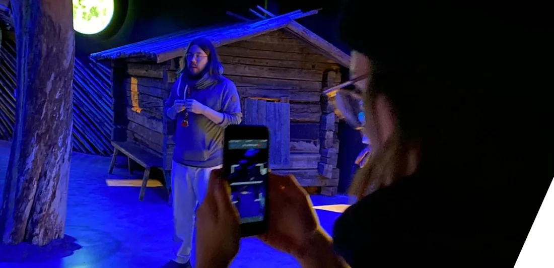 Someone seen from behind films a person speaking on a stage who stands in front of a wooden hut