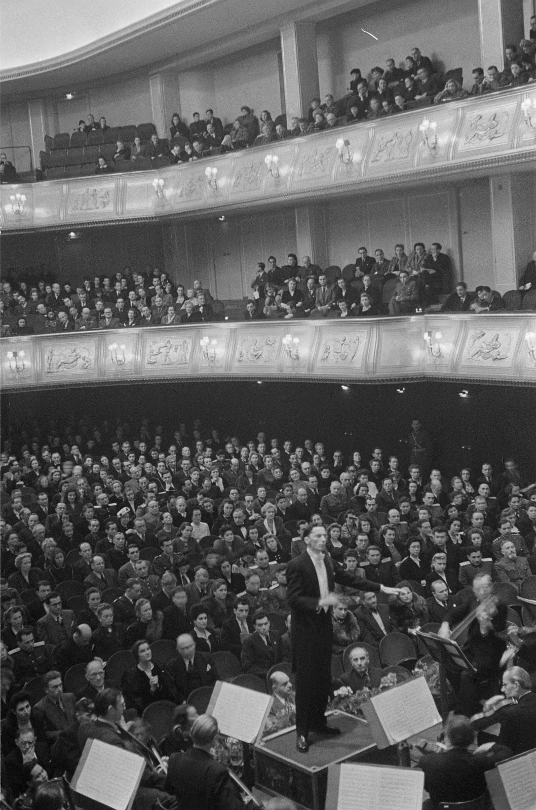 black and white photograph, a conductor and orchestra in a busy concert hall