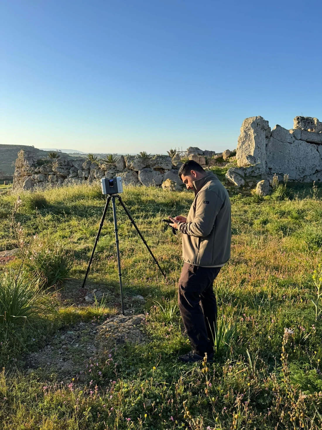 A man stands in a grassy field using equipment on a tripod, with ancient ruins in the background.