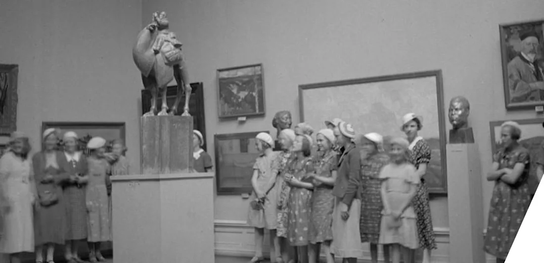 A group of young girls and women in dresses and hats stand in an art gallery, observing a sculpture of a camel with a rider on a pedestal. Various framed paintings and a bust sculpture decorate the walls.