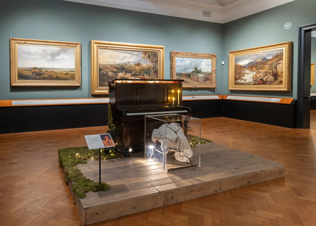 Installation shot of Taylor Swift Songbook Trail at the V&A. A cardigan in a glass box and piano in a room full of paintings. 