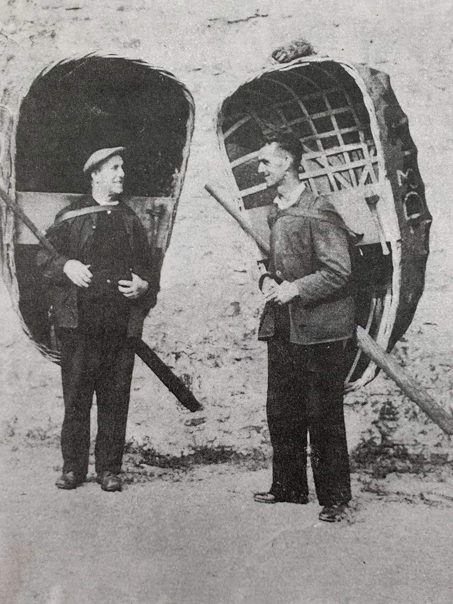 black and white photograph of two standing men, carrying coracle fishing boats on their backs