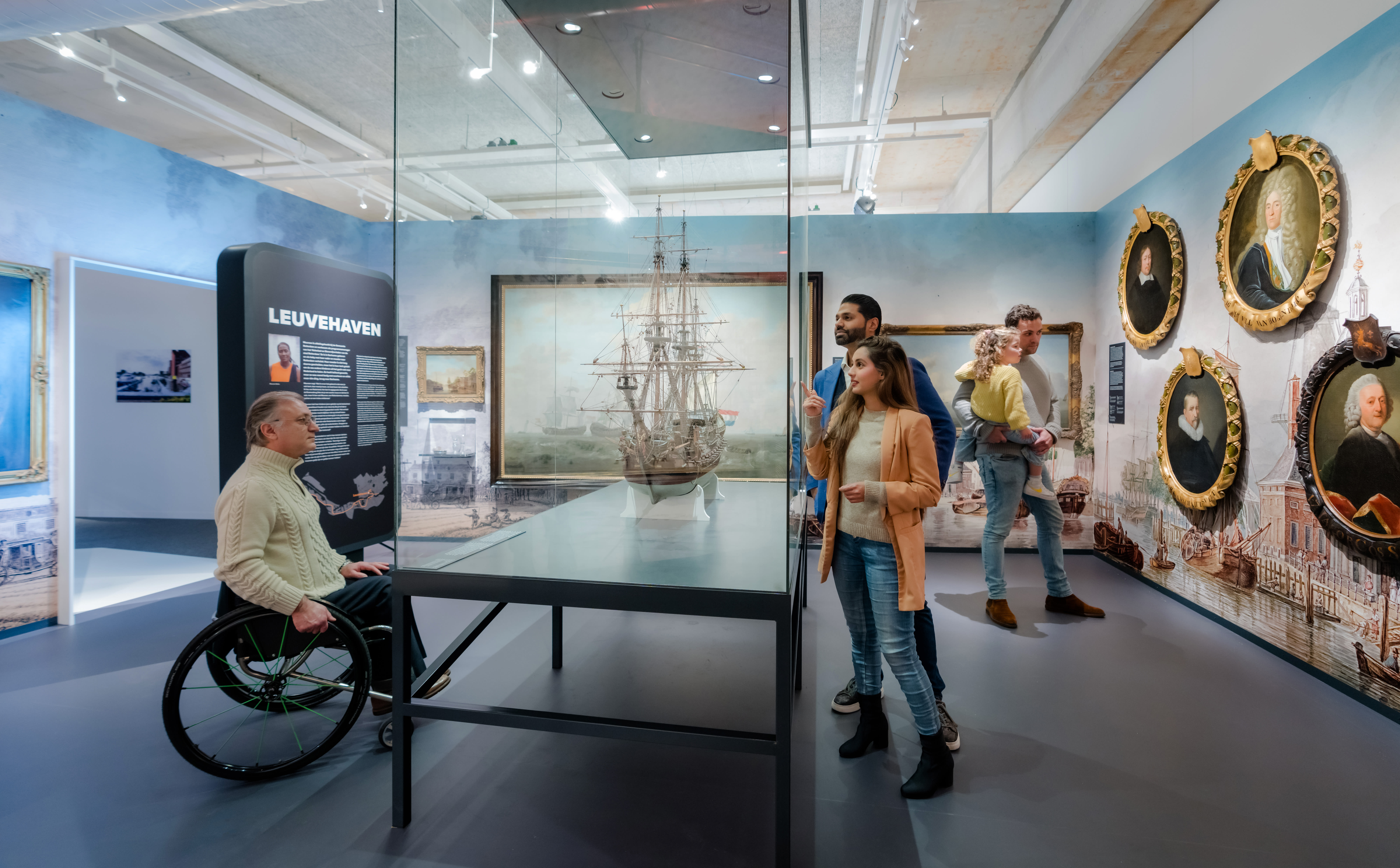 Visitors to the Maritime Museum, including one wheelchair user, look at a model ship in a glass case. Behind them are several portraits and an information board with the title Leuvehaven.