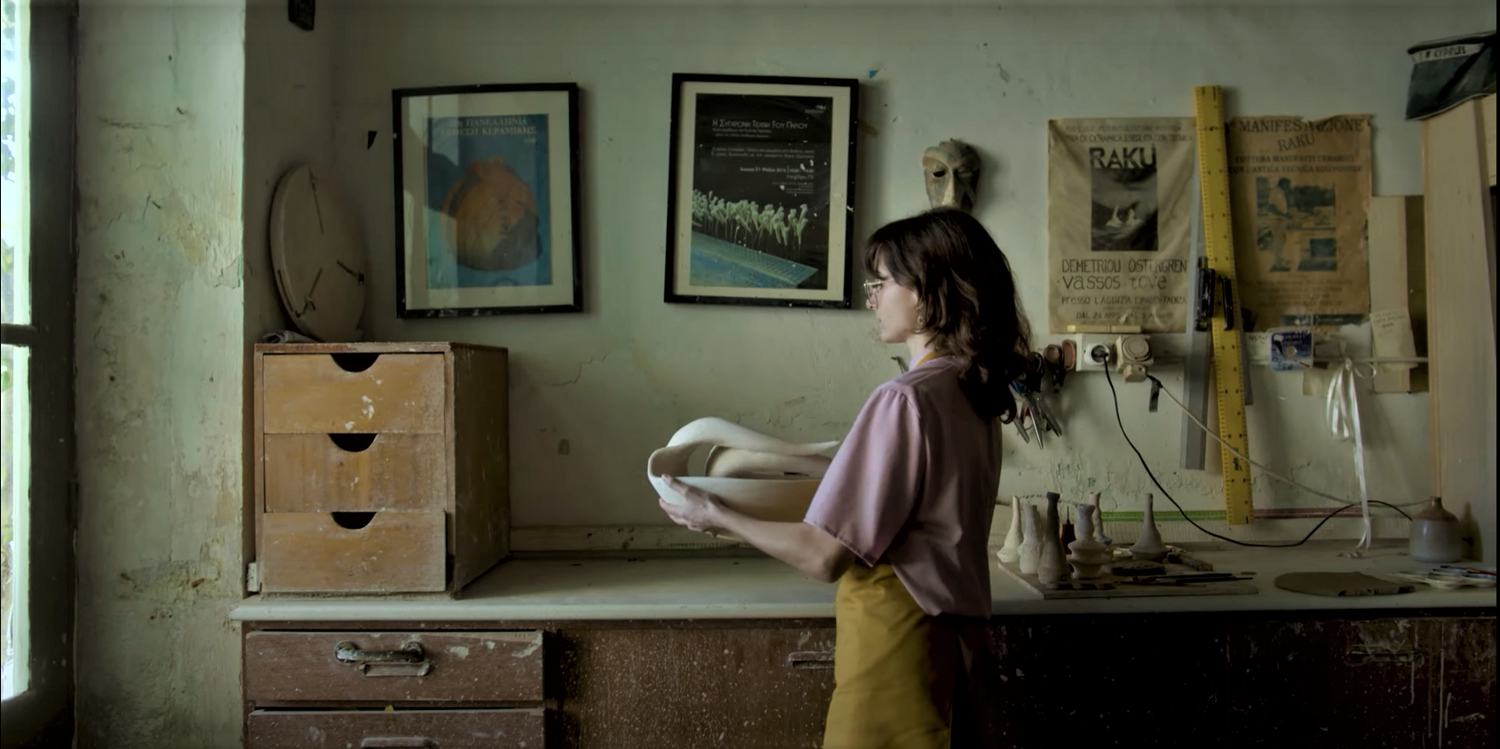 a woman dressed in a pink top and ochre-coloured apron carries an unfired clay sculpture to a workbench in a quaint workshop, light filtering in through windows on the left.