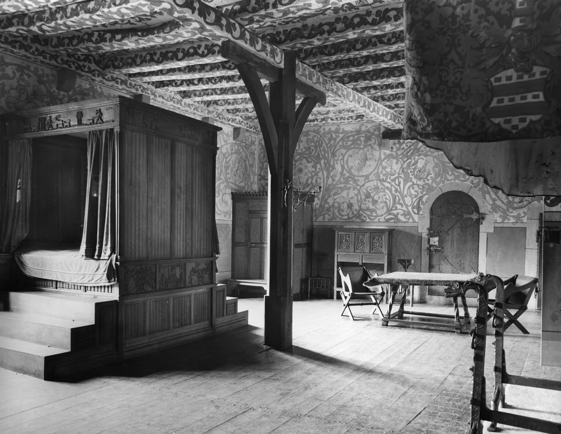 black and white photograph of a room in a castle with a four-poster bed and other furniture such as table, wardrobe, chest 