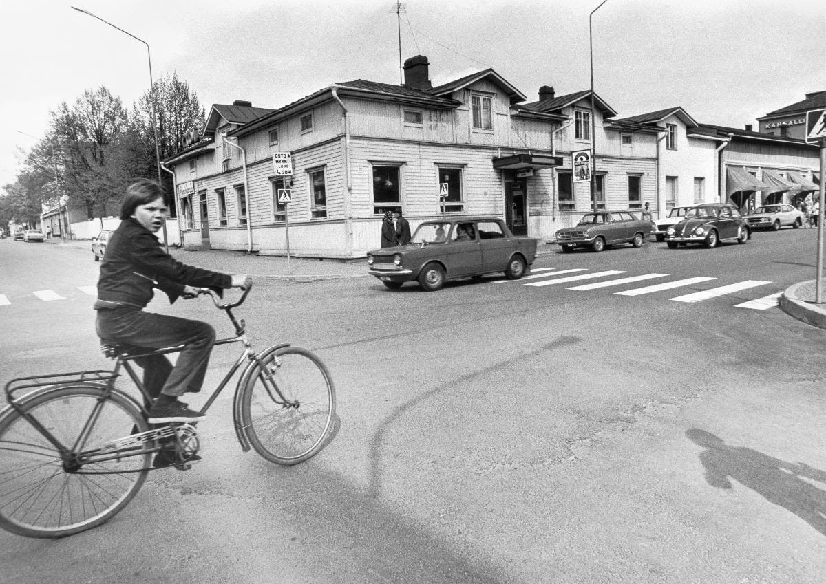 Maneli, men’s meeting place in Heinola, was located on the corner of Torikatu street and Kauppakatu street in Heinola, Finland. Women bought newly-baked bread on the other side of the building in the Maneli Bakery. A boy rides a bicycle along Kauppakatu street.