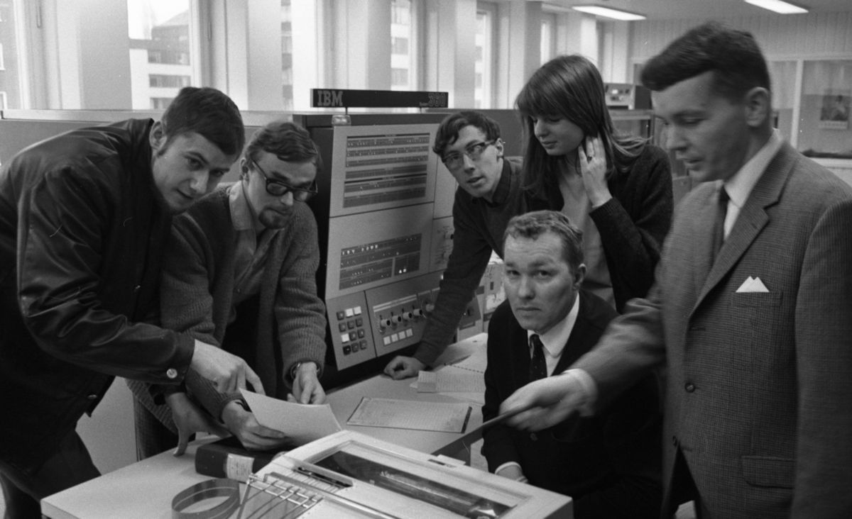 black and white photograph, a number of people in an office looking at equipment.