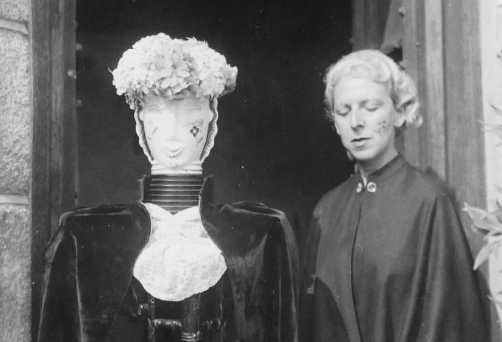 A black and white photograph with a mannequin dressed in black and wearing a white wig next to Claude Cahun, also dressed in a black robe and a white wig. Cahun's eyes are closed.
