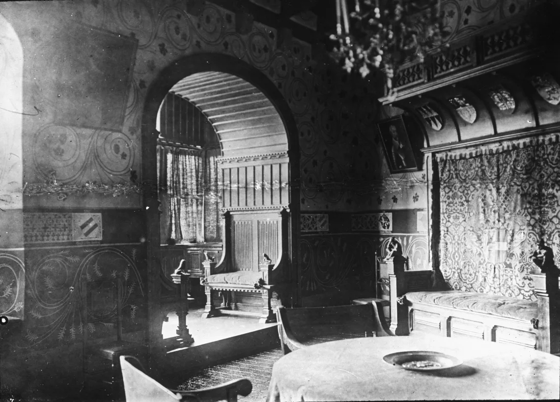 black and white photograph of a room in a castle with a hanging tapestry, table, and other furniture