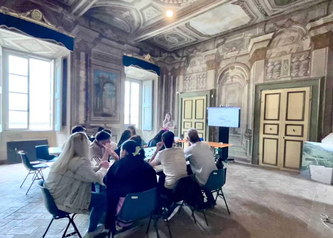 People sit around a table in a grand, ornate room with large windows, listening to a presenter.