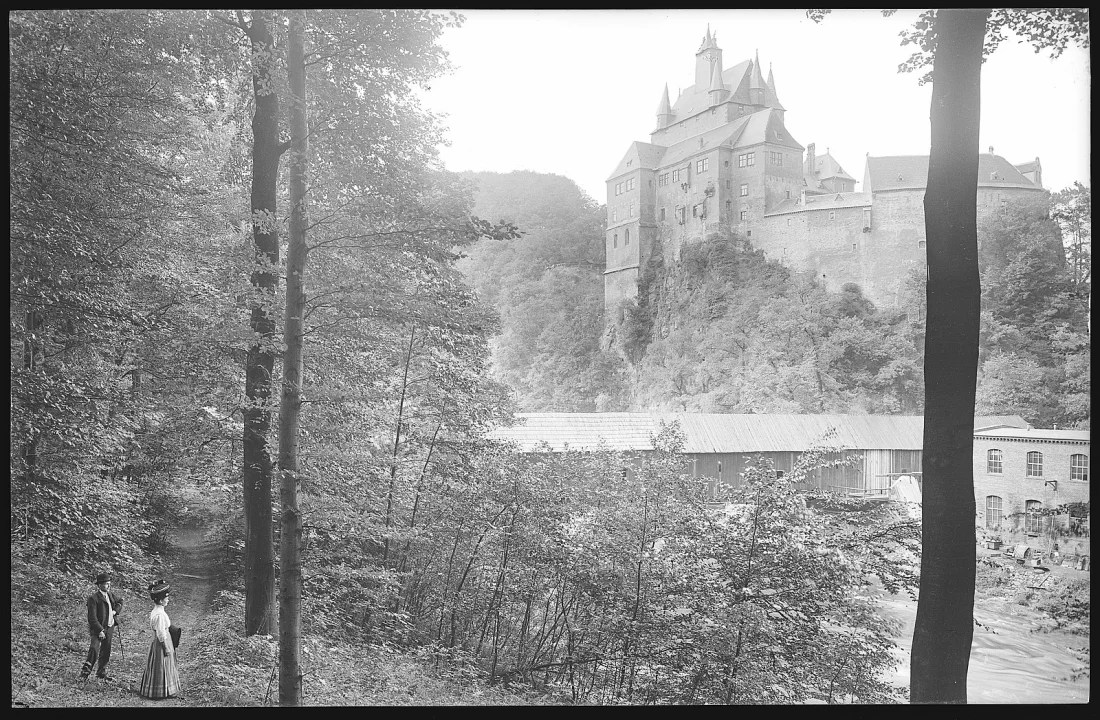 black and white photograph of a castle in the distance with a forest in the foreground where two people stand
