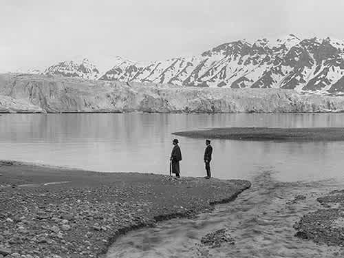 černobílá fotografie dvou lidí stojících před velkým ledovcem