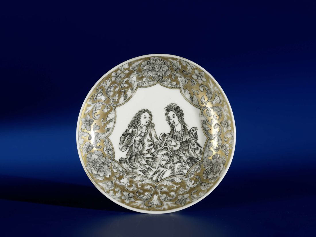 a richly decorated tea saucer standing upright so it shows the glazing decoration of two people in eighteenth-century clothing in a panel of scalloped decoration