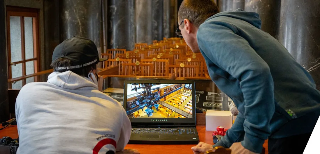 Two people looking at a computer screen on a table in front of a row of chairs and pillars in the background.