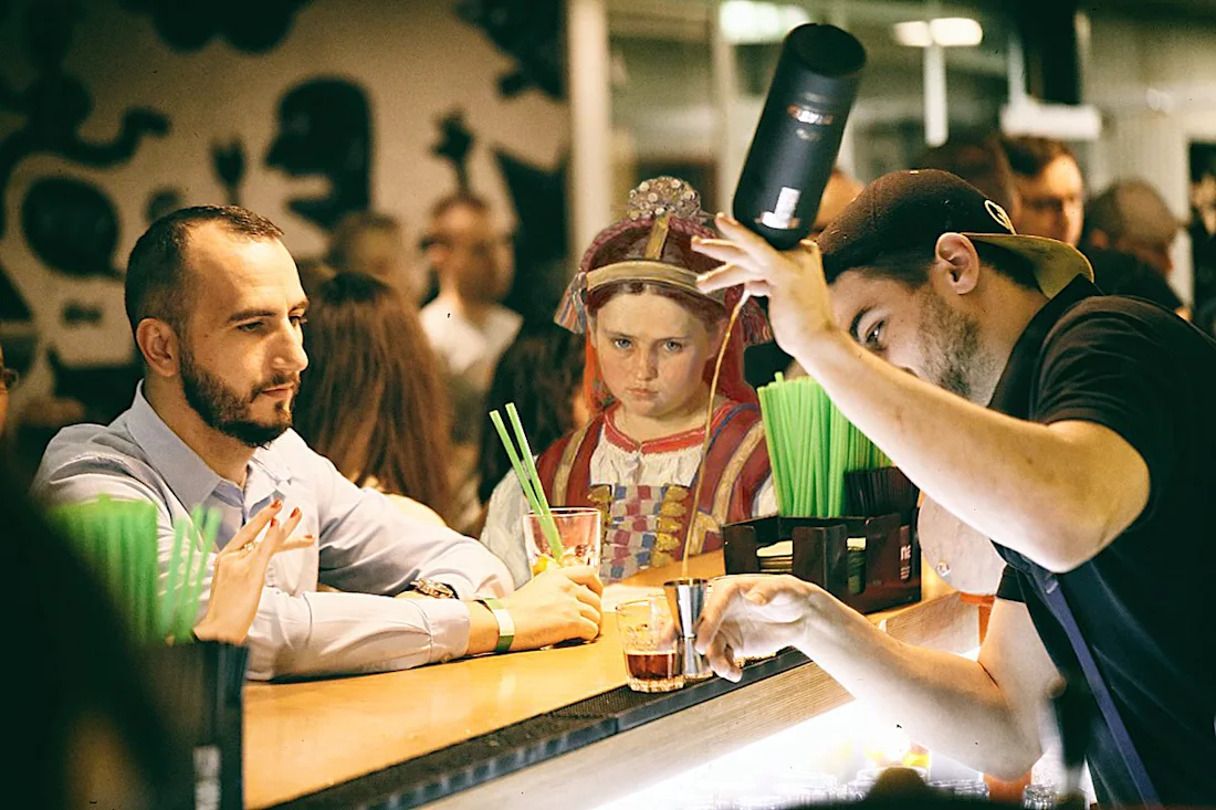A bartender prepares drinks as a man and a woman in historical costume sit at a busy bar.