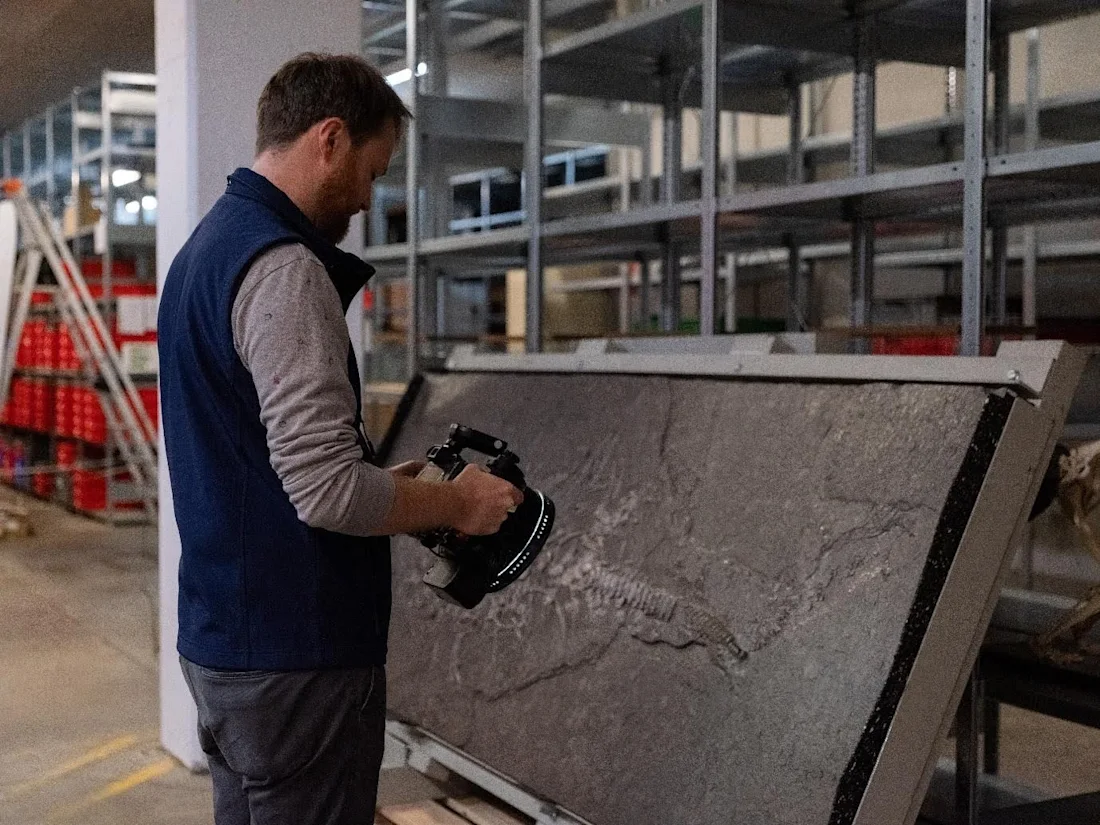 Person examining a large rock slab with handheld equipment in an industrial storage area.