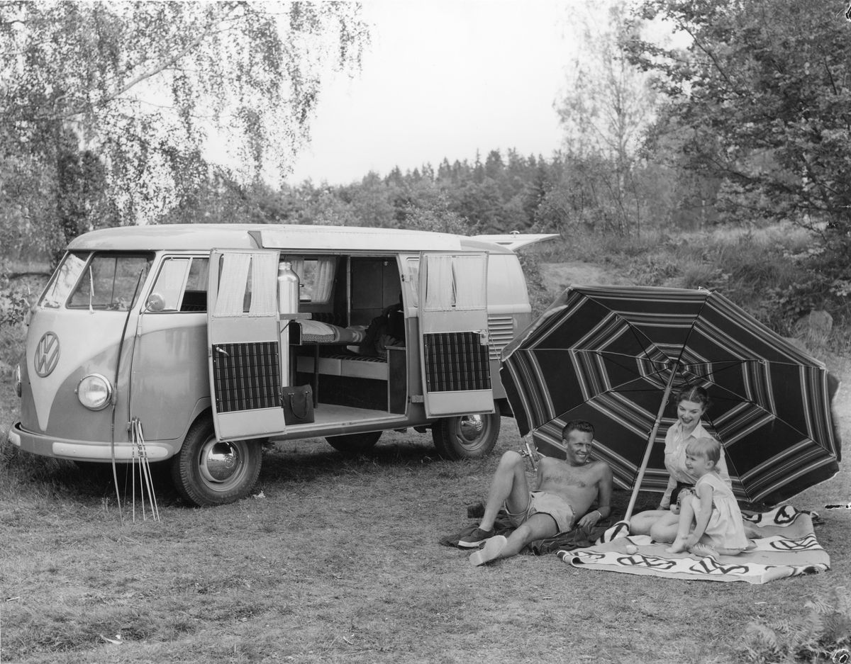 A man, woman and child sit on grass and a picnic blanket, beneath a striped parasol, beside a VW campervan with its doors open. They are surrounded by trees.