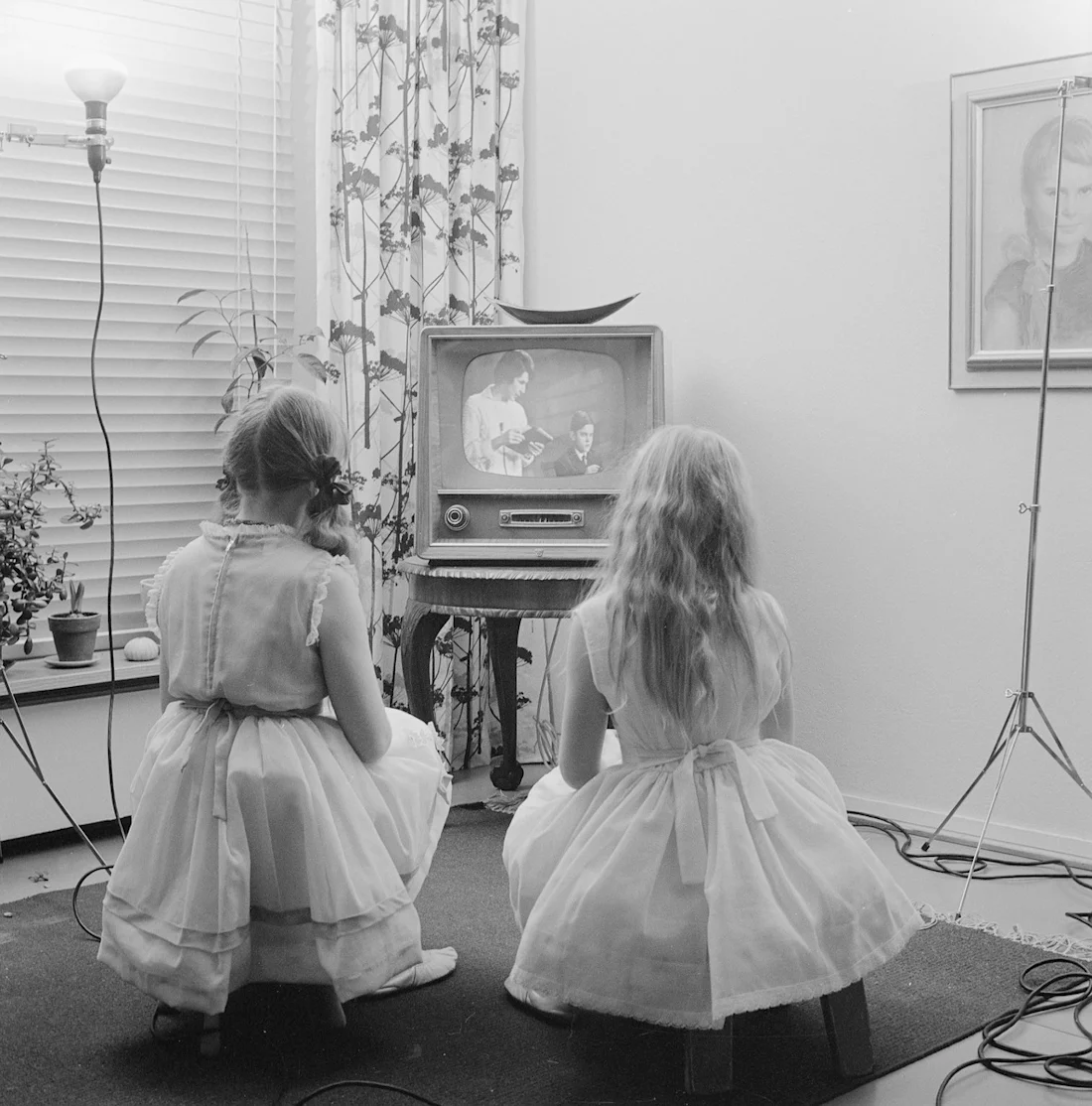 Two girls in dresses sit on stools watching a vintage TV in a living room with plants and framed art.