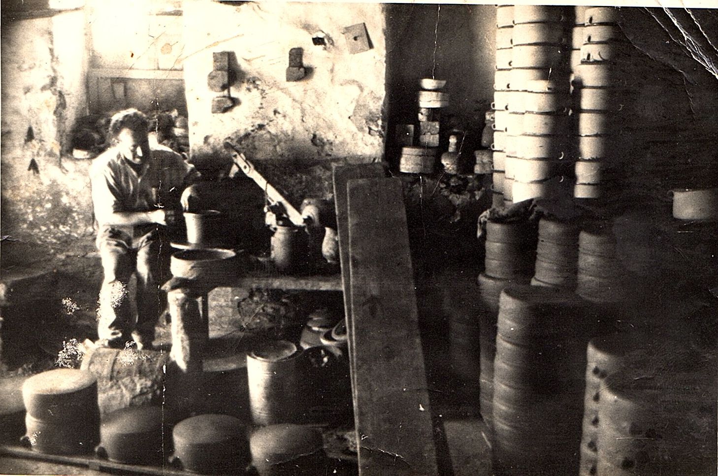 a sepia photograph of a pottery craftsman turning a clay bowl on a turntable. He is surrounded by towering stacks of pottery.