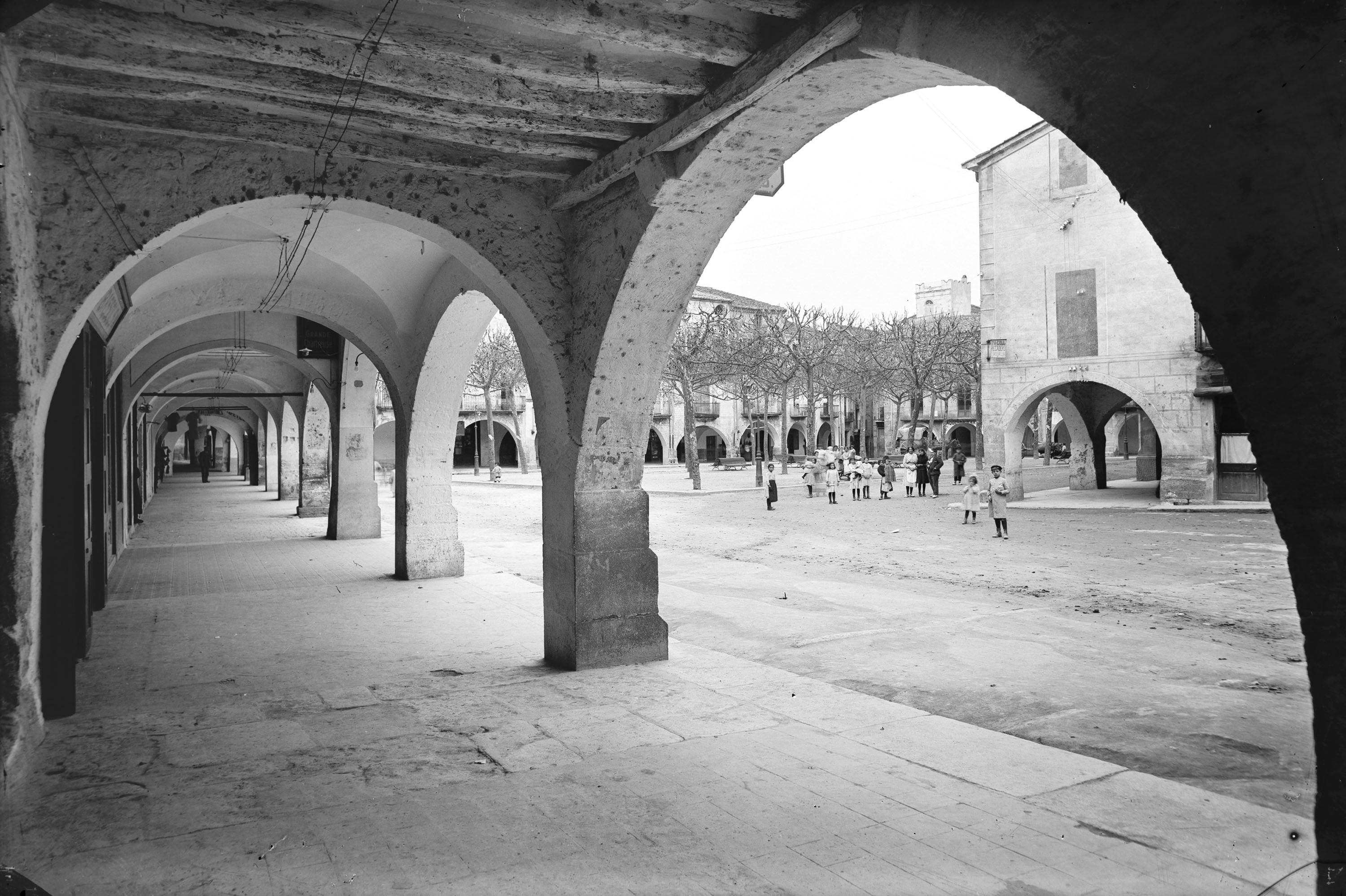 Arched walkway with stone columns leads to an open square where children play, surrounded by trees and buildings.