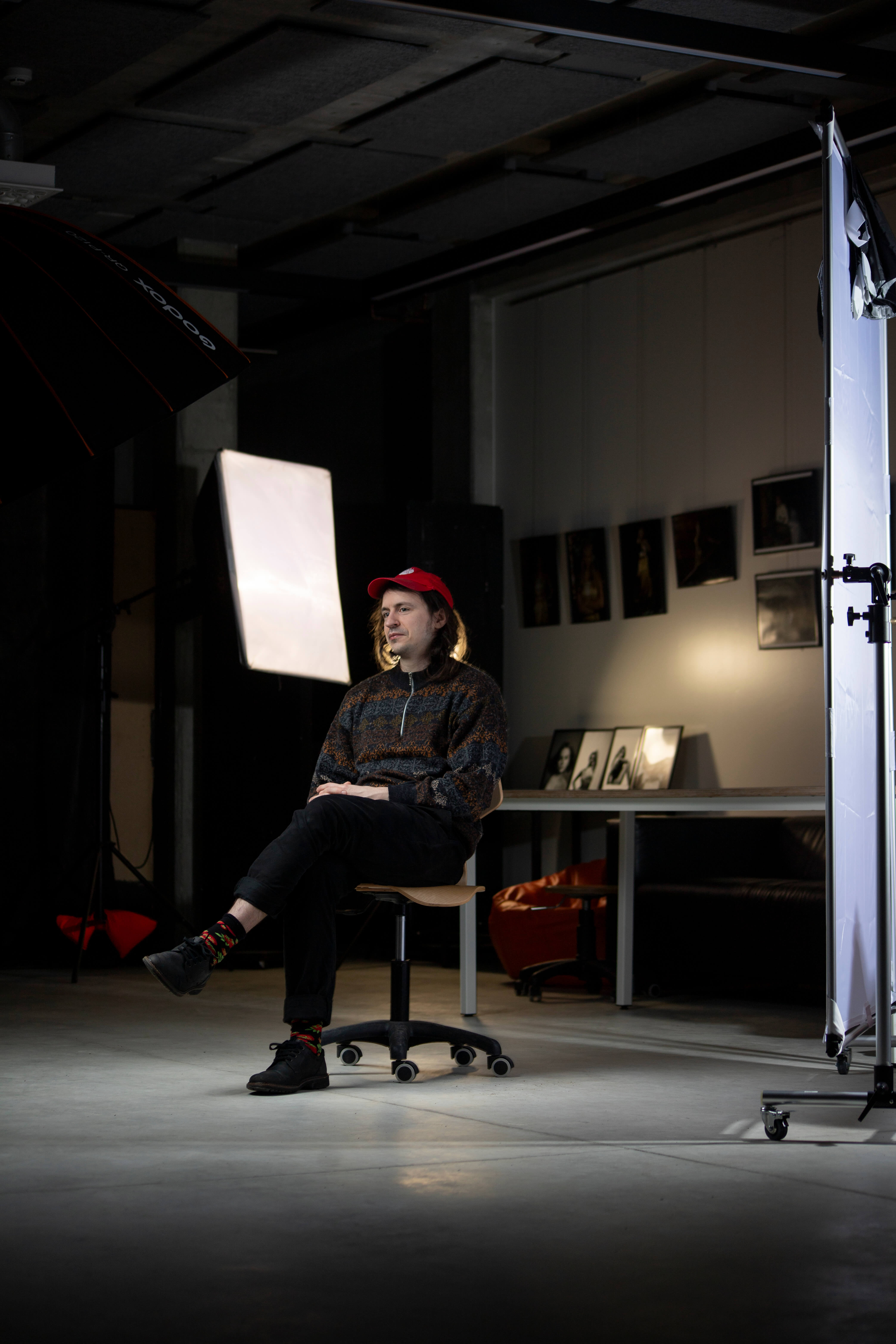 Jānis Jankevics sits on a swivel chair, looking to the left of the frame in a room set up for photography. He wears a red baseball cap, a zip up sweater, dark trousers and dark shoes.