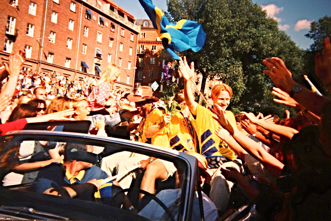 Photographie en couleur, des footballeurs portant des t-shirts jaunes dans une voiture à toit ouvert, agitant un drapeau suédois tandis que la foule tout autour fait la fête.
