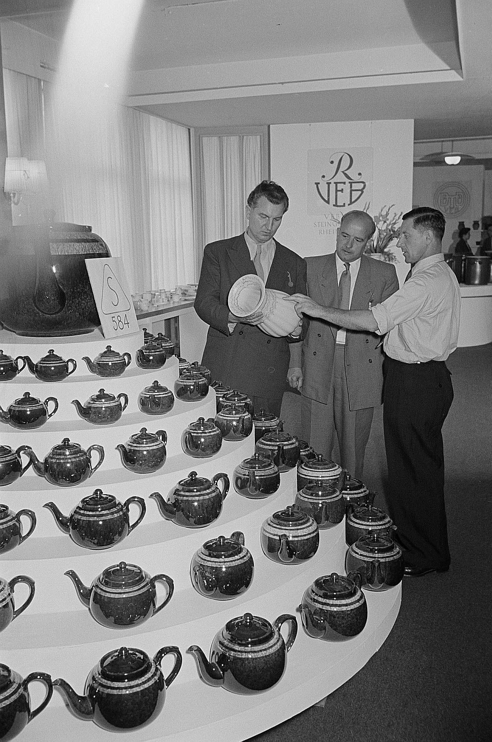 three men admire earthenware, standing in front of a table with dozens of versions of the same teapot placed down next to each other.