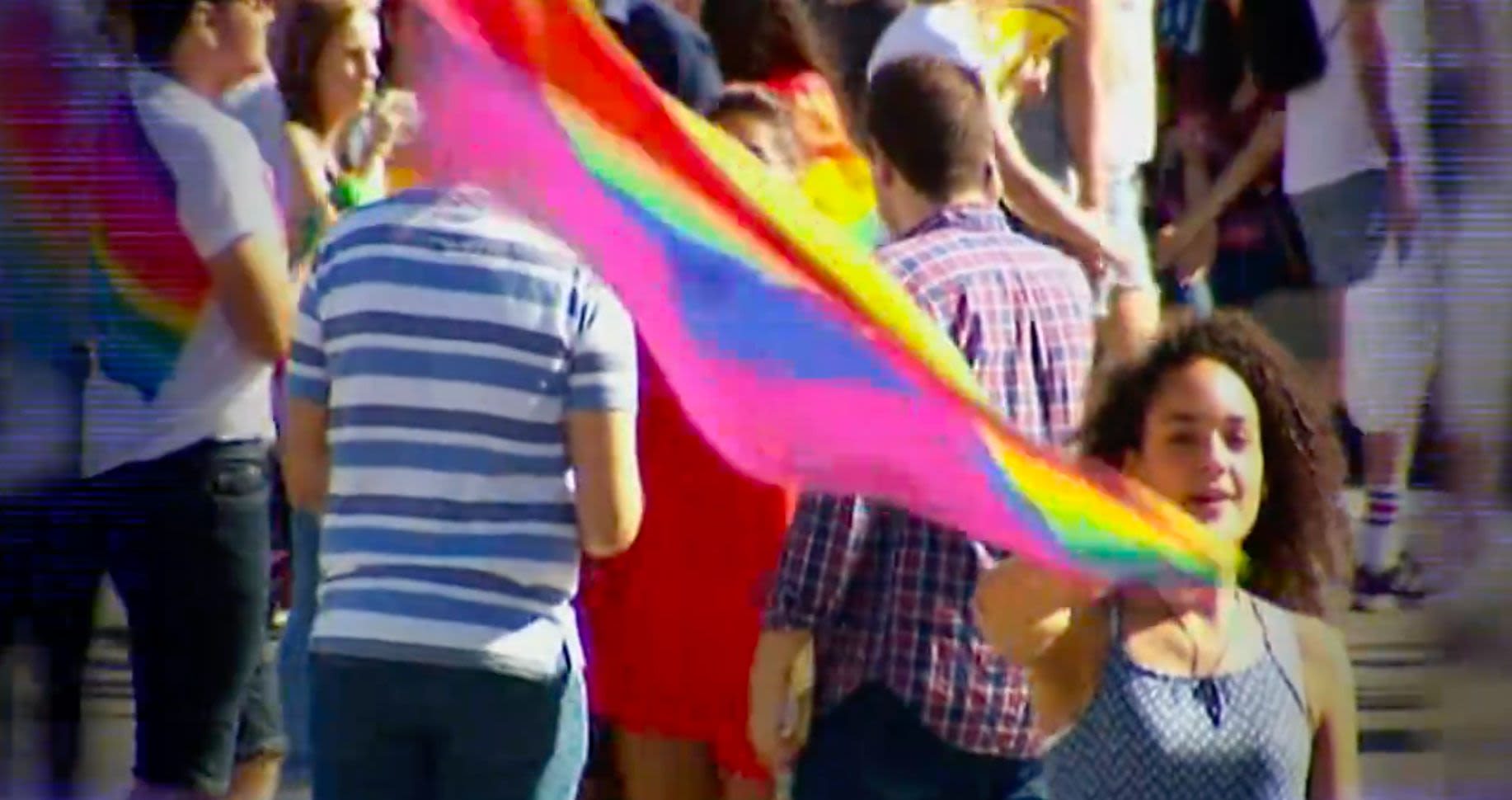 In a crowd of people, a woman with curly hair is waving the pride flag. 