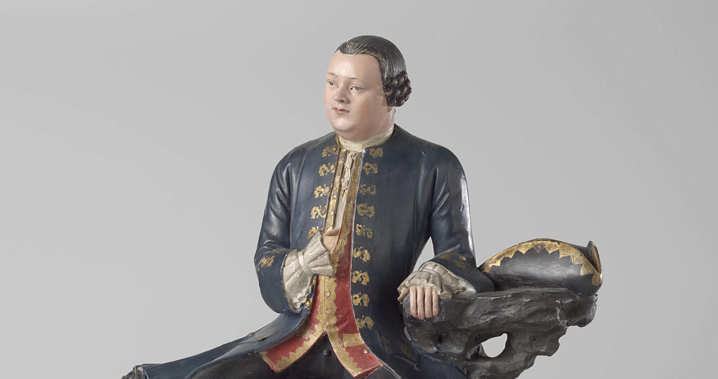 a porcelain figure of a man sitting down, wearing a long blue jacket with gold trim and gold tassels, his hair slicked back and ending in Victorian-style ruffles