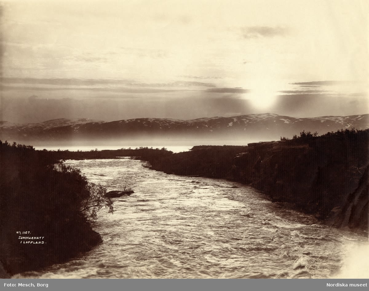 A  black and white postcard of a river, and snowy mountains, with the sun low in the sky. 