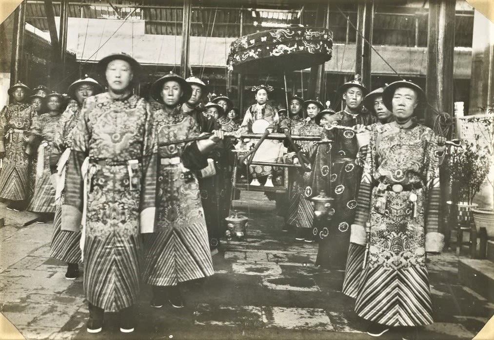 Black-and-white photograph. A group of Chinese servants and officials pose in ornate clothing, with the empress Cixi in a wooden carrier in the middle of the frame.