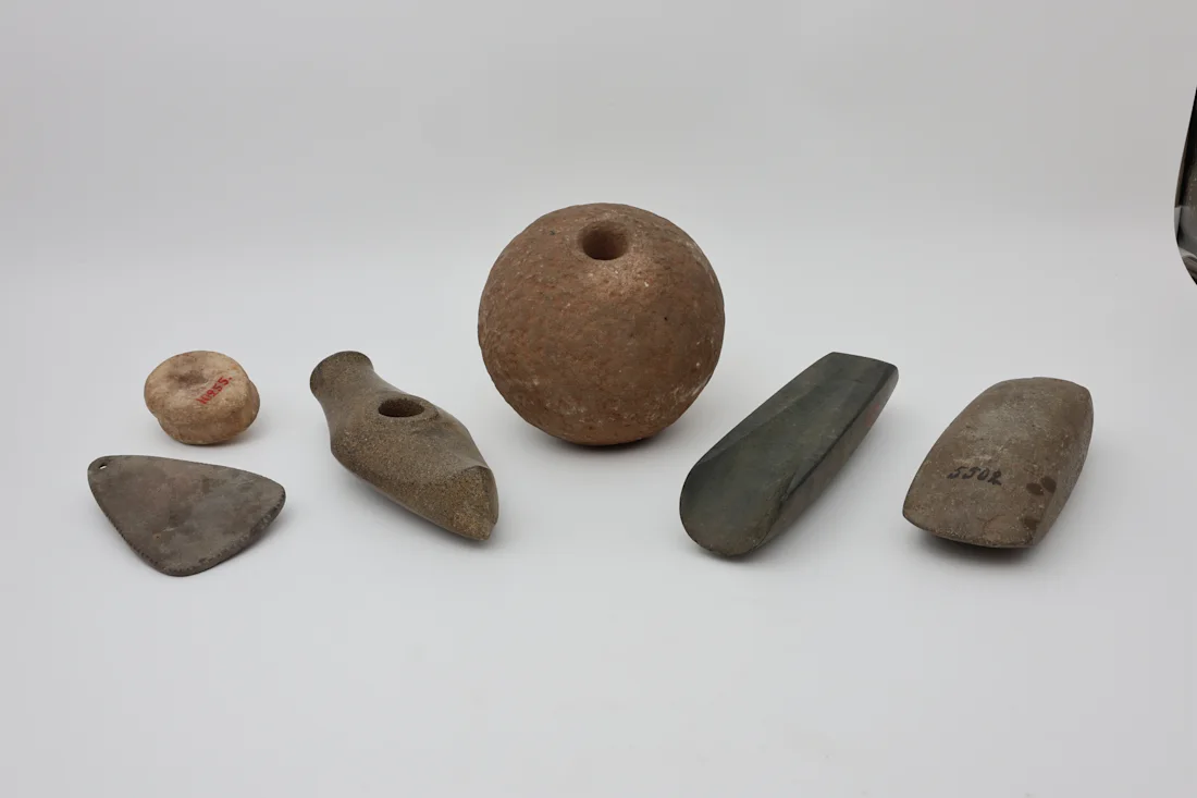 Six ancient stone tools and artifacts arranged on a white background, including axes and a perforated sphere.