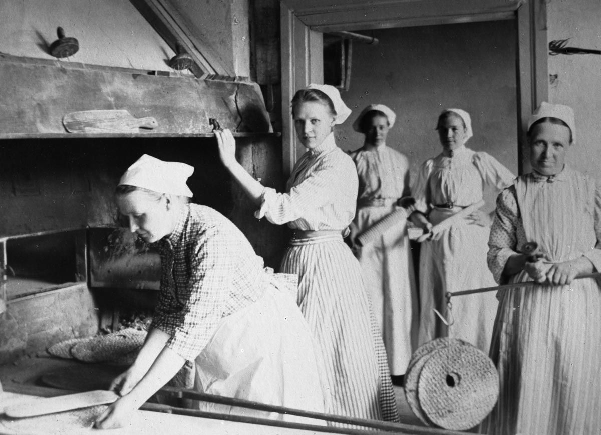 a black and white photograph, five women standing by a large open oven.