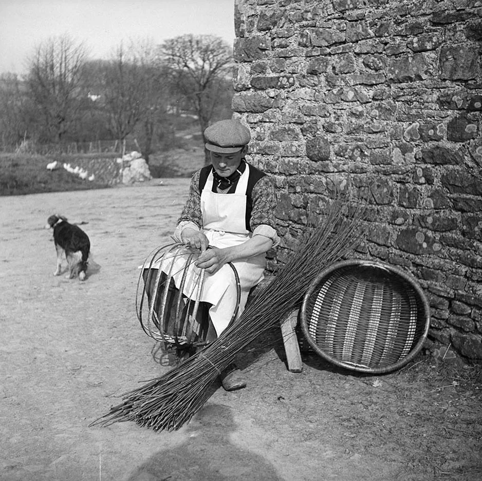 black and white photograph of a man sitting outside weaving a basket, a dog in the background