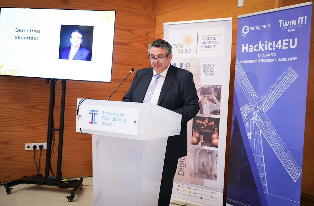 A man in a suit speaks at a podium with banners and a screen behind him.