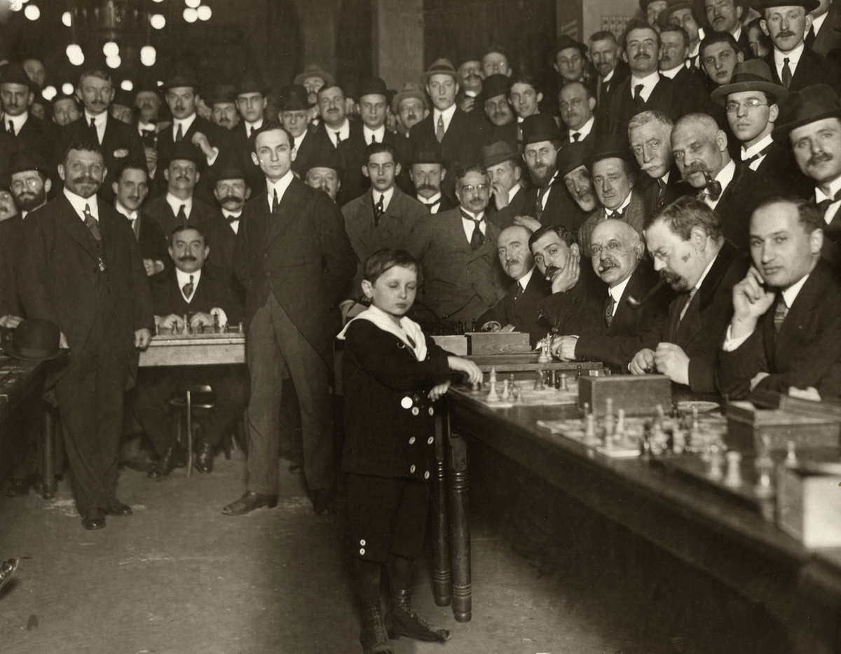 black and white photograph, a young boy playing chess with a large crowd of men looking on.