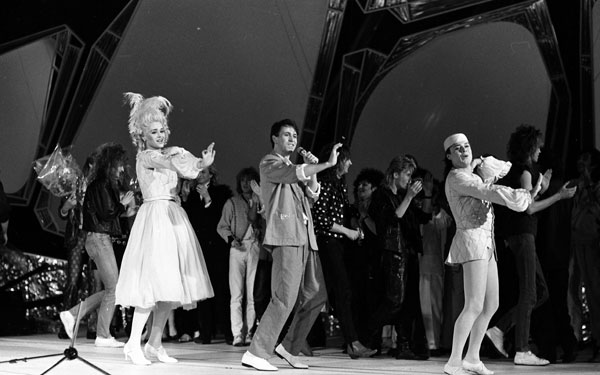 black and white photograph of three people singing and dancing on stage with people standing behind them