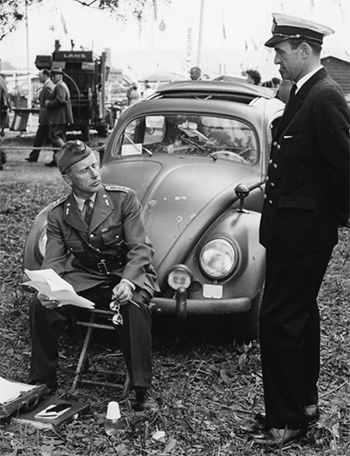 two men in military uniform, one standing, one sitting, in front of a Volkswagen beetle