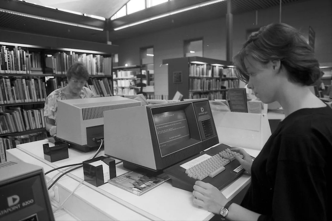Two people use vintage computers in a library, surrounded by bookshelves and stacks of books.